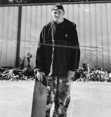 Black and white image of a young man standing with a skateboard, wearing a cap and a casual jacket. He is in front of a fence with some greenery in the background, dressed in camouflage pants.
